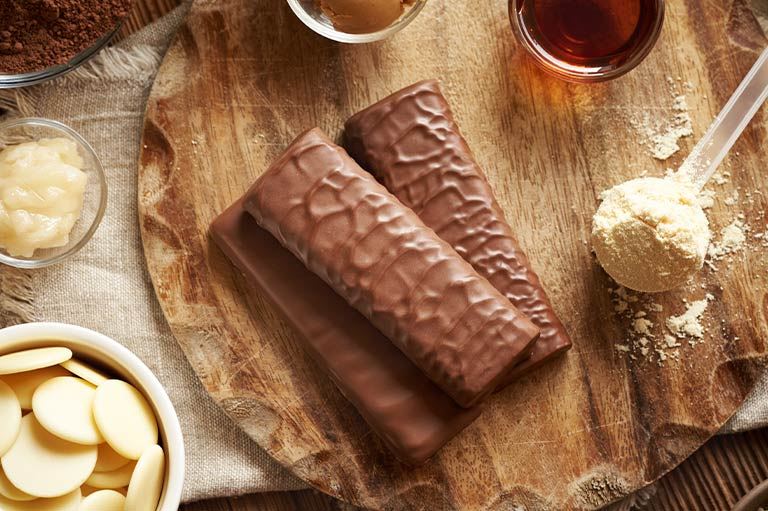 Tres barras de proteína Pro-TF de chocolate y crema de maní y una cuchara de proteína de vainilla en una tabla de madera.
