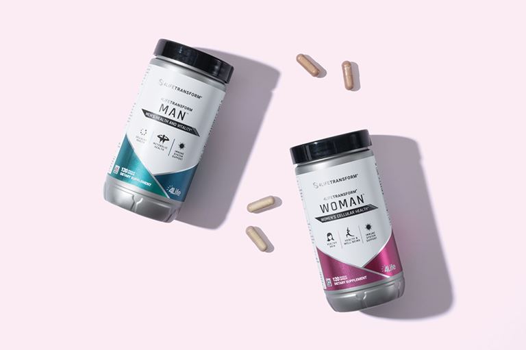 Una botella de 4Life Transform Man y otra de Transform Woman sobre una superficie plana de color rosa claro, con un par de pastillas cerca de las botellas.