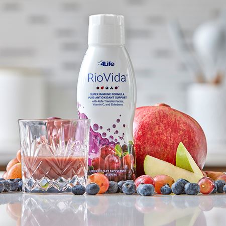 Bottle of RioVida on a flat surface with a half-filled cup of juice, a pomegranate, apple slices, blueberries, and açaí.